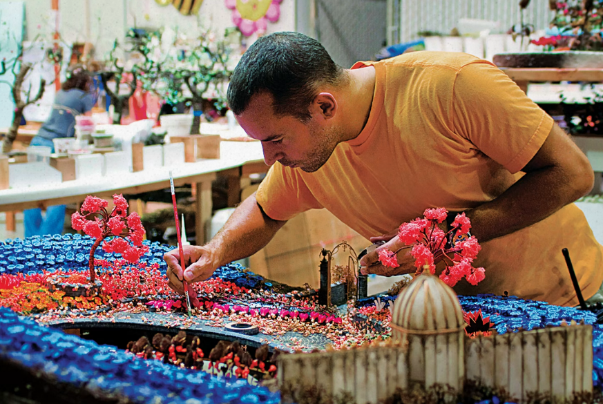 A man with tan skin and dark close-cropped hair leans over a large garden diorama from the movie Coraline with blue, pink, and orange flowers as he paints some of the ground by the flowers.